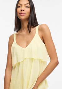 Vrouw met lang donker haar die een mouwloze, lichtgele gestapelde top draagt, zacht glimlachend tegen een effen witte achtergrond.