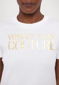 White cotton T-shirt with gold metallic print reading "VERSACE JEANS COUTURE." Short sleeves and crew neckline. Smooth texture.