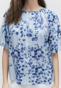 Lyseblå kortærmet bluse med et blødt blomstermønster i mørkere blå nuancer, med en nøglehulsudskæring og afslappet pasform.