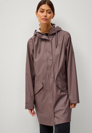 Kobieta w długim, mauve przeciwdeszczowym płaszczu z kapturem, zapinanym na zatrzaski, z przednimi kieszeniami z klapkami, stojąca na tle jednolitego jasnoszarego tła.