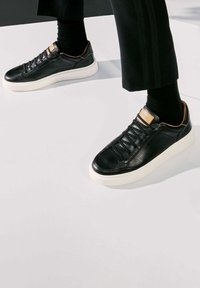 Baskets en cuir noir avec un panneau latéral texturé, semelle blanche et accents beige au talon et à la languette ; elles comportent un laçage traditionnel.