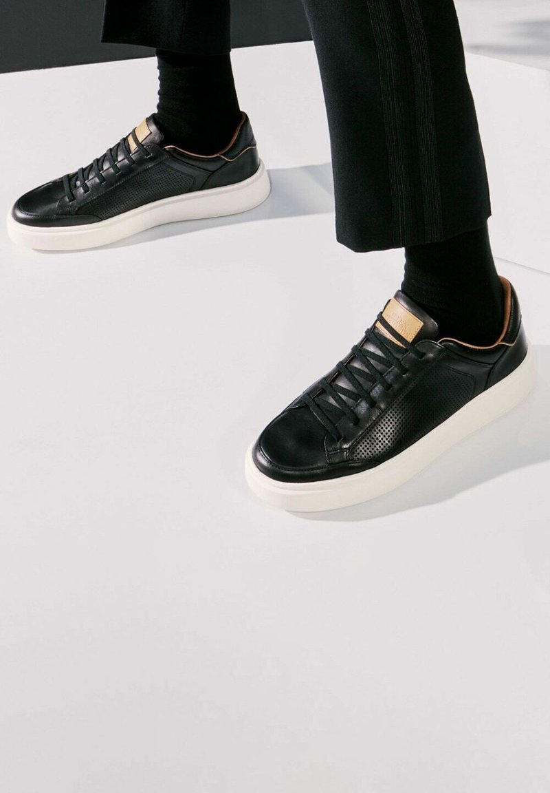 Baskets en cuir noir avec un panneau latéral texturé, semelle blanche et accents beige au talon et à la languette ; elles comportent un laçage traditionnel.