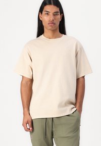 Beige katoenen T-shirt met korte mouwen, ronde hals en een subtiele geborduurde logo op de borst, gedragen met olijfgroene broek.
