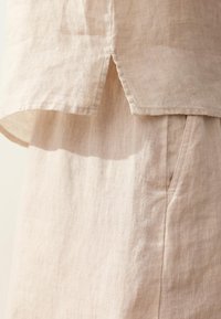 Tissu en lin beige présentant une fente latérale sur un haut ample superposé à un pantalon assorti avec une couture de poche visible.