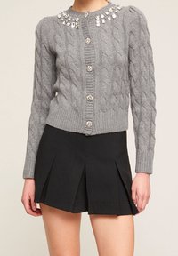 Šedý pletený cardigan s kabelkou, s proužkovaným límcem a velkými ozdobami, zapínáním na knoflíky a přiléhavými manžetami, v kombinaci s skládací černou sukní.