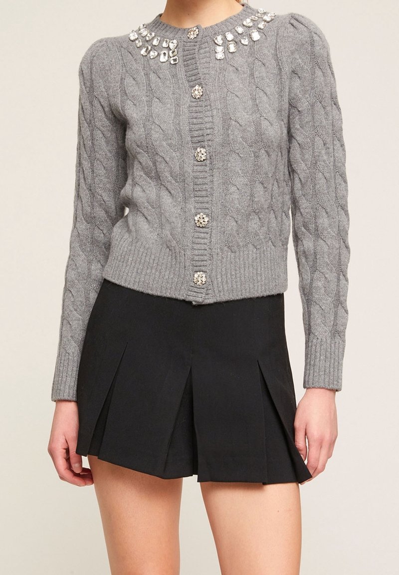 Šedý pletený cardigan s kabelkou, s proužkovaným límcem a velkými ozdobami, zapínáním na knoflíky a přiléhavými manžetami, v kombinaci s skládací černou sukní.