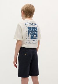 T-shirt crema con stampa grafica blu e testo sul retro; pantaloni navy con tasche posteriori e dettagli a bottone. Bambino in piedi, visto da dietro.