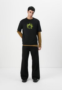 Black t-shirt with green sun and smiley graphic, layered over a brown long-sleeve shirt, paired with black wide-leg pants and black shoes.