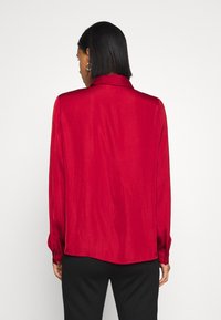 Červená blůza s dlouhým rukávem vyrobená z hladkého, lesklého materiálu s špičatým límcem a knoflíkovými manžetami. Má uvolněný střih a jemné splývání.