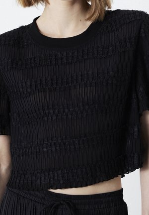 Frau trägt ein schwarzes, strukturiertes Crop-Top mit horizontalen Streifen und passende plissierte Hosen mit Kordelzug in der Taille.