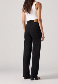 Femme aux longs cheveux portant un haut blanc sans manches et un jean noir taille haute à jambe droite, debout, de dos sur un fond clair.