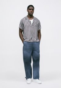 Uomo in piedi con le mani nelle tasche, che indossa sneakers bianche, pantaloni blu, una maglietta bianca e una camicia a maniche corte a quadri su uno sfondo bianco.