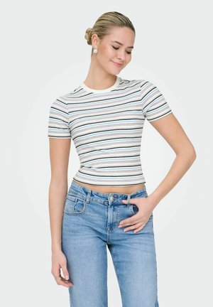 Young woman with blonde hair in a bun wearing silver earrings, a short-sleeved striped crop top, and light blue jeans against a white background.
