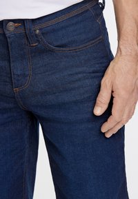 Jeans de mezclilla en azul oscuro con una textura suave, que presentan costuras naranjas, un bolsillo frontal y un cierre de botón metálico.