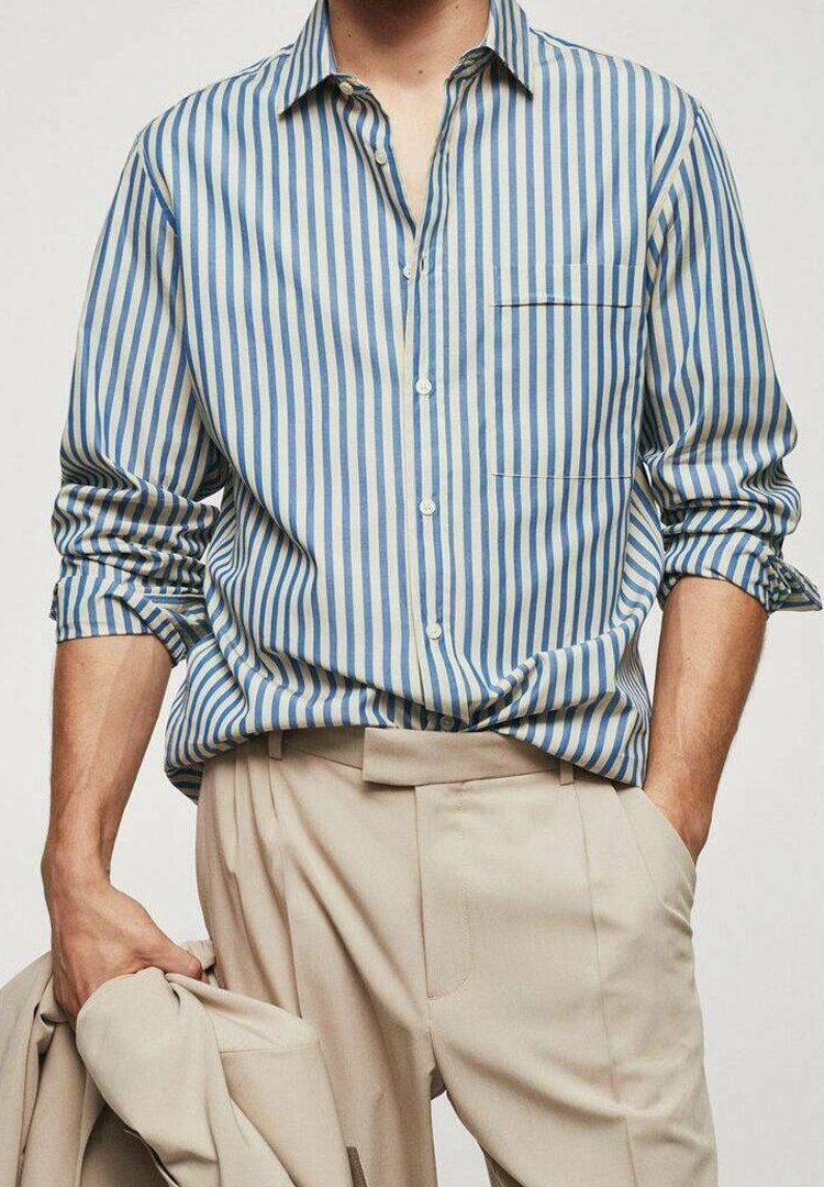 Chemise à rayures bleues et crème avec boutons et poche poitrine. Manches longues retroussées aux poignets, portée avec un pantalon beige.