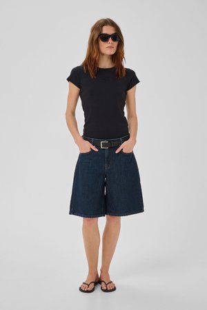 Kobieta ubrana w czarny t-shirt, ciemne długie szorty jeansowe z paskiem, czarne okulary przeciwsłoneczne oraz czarne japonki, stoi z rękami w kieszeniach na szarym tle.