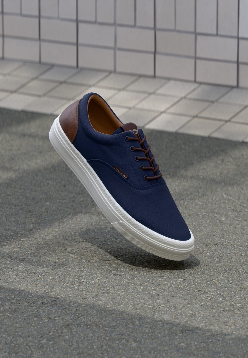 Navy canvas sneaker with brown leather accent and white rubber sole, featuring round laces and a smooth texture.