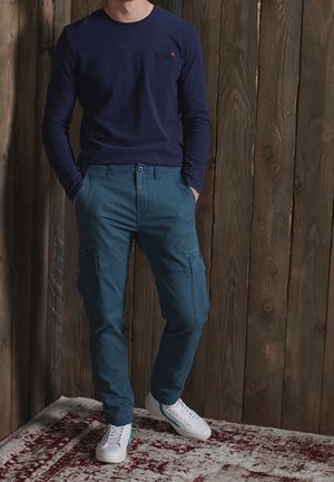 Uomo in piedi con le mani in tasca che indossa una maglia blu navy a maniche lunghe, pantaloni cargo verde acqua e scarpe da ginnastica bianche su un tappeto a motivo.