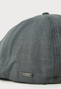 Casquette grise en tissu texturé, de forme arrondie avec une étiquette en métal argenté attachée sur le côté.