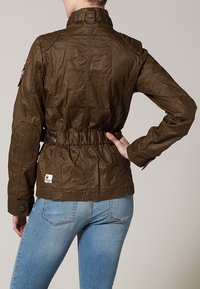 Brun, texturerad jacka med hög krage, åtdragen midja och detaljerad sömnad. Har axelplattor och knappar som dekoration.