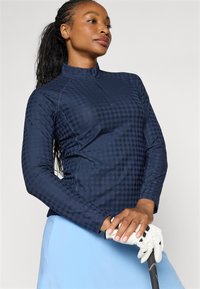 Marineblå quarter-zip genser med hundetannmønster, lange ermer og strukturert stoff. Modellen holder en golfkølle og har på seg et lyseblått skjørt.
