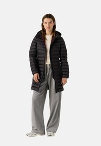 Abrigo largo acolchado negro con capucha y bolsillos laterales. Presenta un diseño de acolchado texturizado. Llevado sobre un suéter ligero y pantalones grises.