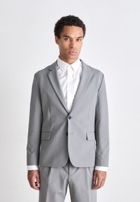 Chaqueta de traje gris ajustada, con cierre de un botón, solapas con muesca, bolsillos laterales, tejido de textura suave, combinada con una camisa blanca.