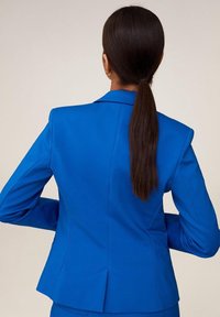 Žena s dlouhými tmavými vlasy staženými do nízkého culíku, oblečená v jasně modrém přiléhavém blejzru, stojící s rukama v bok proti jednobarevnému pozadí.