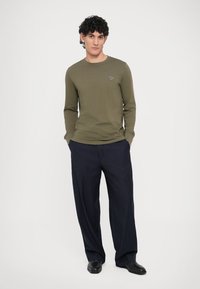 Olīvu zaļa garroku krekls ar apaļu kakla izgriezumu un mazu emblēmu. Saskaņots ar tumšām plaša piegriezuma biksēm ar smalkām svītrām un melniem apaviem.