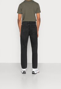 Vaqueros de mezclilla negros con corte recto, cinco bolsillos y un sutil logotipo rojo en el bolsillo trasero. Combinados con una camiseta verde oscura y zapatillas blancas.