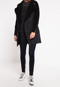 Cappotto in pelle ecologica nera con un ampio colletto e texture in shearling, indossato sopra un maglione scuro e jeans scuri scintillanti, abbinato a sneaker argentate.