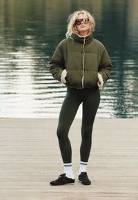 Oliwkowo-zielona puchowa kurtka z podszyciem z baranka, zestawiona z ciemnozielonymi legginsami i czarnymi trampkami. Całość dopełniają odblaskowe okulary przeciwsłoneczne.