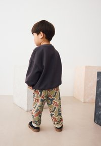 Enfant portant un sweat-shirt gris foncé ample et un pantalon cargo camouflage aux teintes de vert, brun et rouge, assortis à des chaussures noires.