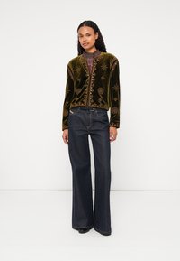 Zöld bársonykabát bonyolult arany hímzéssel, kerek nyakkivágással, gombokkal rögzítve, magas derekú sötét farmernadrággal párosítva.