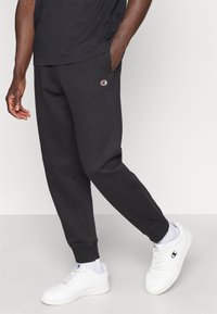 Μαύροι παντελόνες φόρμας κατασκευασμένοι από μαλακό ύφασμα με κωνικό σχέδιο, διακοσμημένοι με μικρό λογότυπο στην αριστερή τσέπη, συνδυασμένοι με λευκά αθλητικά παπούτσια.