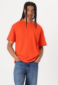 T-shirt orange à col rond avec des manches courtes, fabriqué en coton doux. Le modèle porte un jean en denim bleu, se tenant avec les mains dans les poches.