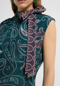 Grünes gemustertes Kleid mit hohem Kragen und einem lila Krawatten-Detail. Der Stoff weist wirbelnde Designs in Creme- und Brauntönen auf.