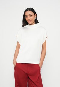 Valkoinen, lyhythihainen paita korkealla kaula-aukolla, rennolla istuvuudella ja pehmeällä, sileällä tekstuurilla. Yhdistetty punaisiin, leveälahkeisiin housuihin.