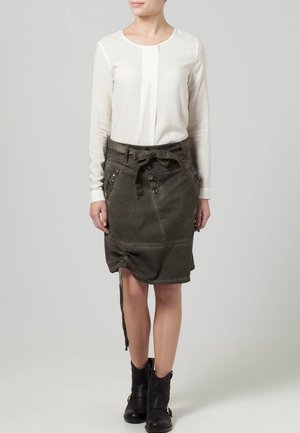 Blouse blanche à manches longues avec un détail plissé subtil, associée à une jupe verte mi-longue munie de boutons et d'un ourlet à cordon, portée avec des bottes noires.