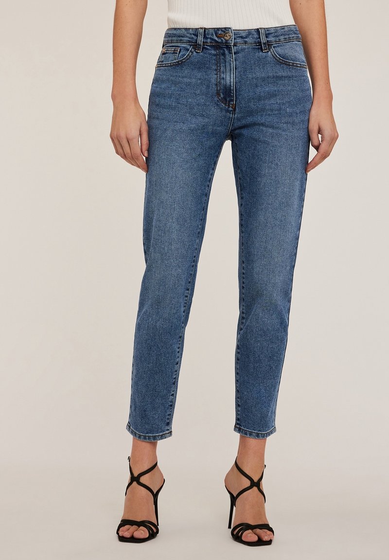 Niebieskie dżinsy z średnim stanem, zwężanymi nogawkami i klasycznym pięciopocketowym wzorem. Noszone z czarnymi sandałami na wysokim obcasie z paskami.