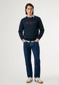 Tumši zils sporta krekls ar sarkanu "Pepe Jeans" logotipu, gaiši zila krekla krekls ar apkakli zem tā, zili džinsa džinsi un baltas sporta kurpes ar melnām detaļām.