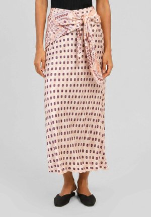 Femme portant une jupe midi rose clair avec un motif carré foncé et un détail d'écharpe assortie nouée à la taille, associée à des chaussures plates noires.