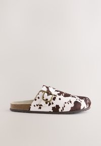 Sabot à enfiler avec motif imprimé vache blanc et brun foncé et une bride à boucle en laiton sur le côté, avec une semelle en liège et une semelle en caoutchouc noire.