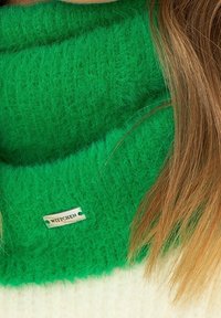 Écharpe verte en tricot duveteux avec une étiquette WITTCHEN. Présente une texture douce et un design large. Des cheveux bruns sont partiellement visibles à côté.