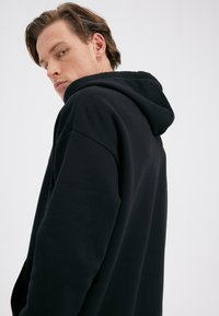 Black hooded sweatshirt with a relaxed fit. Features a kangaroo pocket, ribbed cuffs, and is made from a soft, textured fabric.