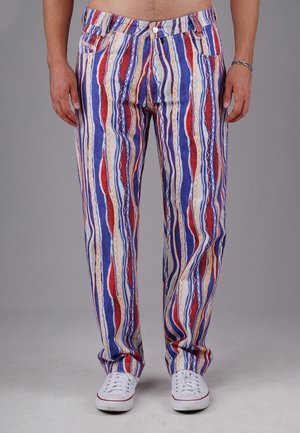 Homme portant un pantalon à rayures verticales rouges, bleues, blanches et beiges avec des baskets blanches, debout devant un fond gris uni.