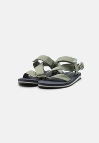 Green fabric sandals with a cross-strap design, dark rubber soles, and adjustable buckle detail, featuring a textured footbed for grip.