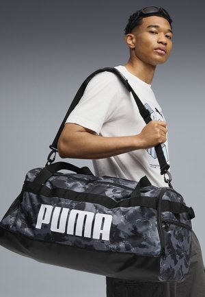 Junger Mann mit Sonnenbrille auf dem Kopf und weißem Grafik-T-Shirt trägt eine schwarze und graue Puma-Tasche mit Camouflage-Muster über der Schulter.