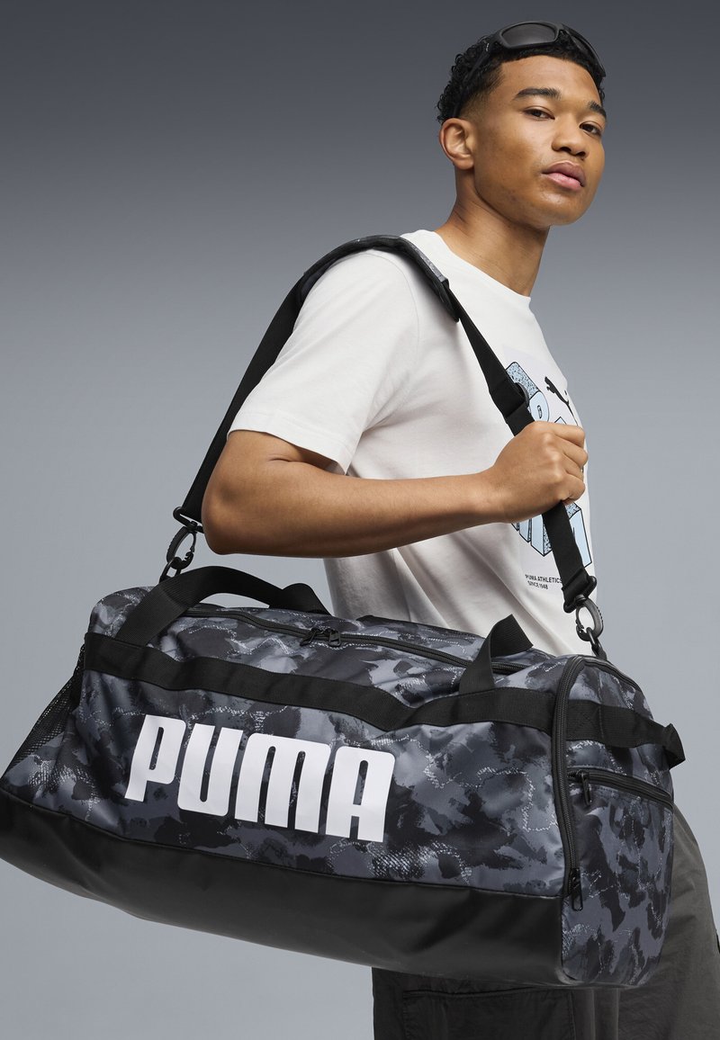 Joven con gafas de sol en la cabeza y camiseta blanca con diseño gráfico lleva un bolso de viaje Puma camuflado en negro y gris sobre el hombro.