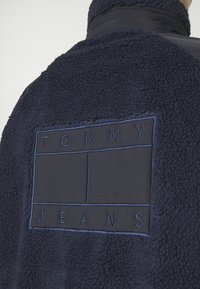 Giacca in fleece blu navy con superficie texturizzata e una patch rettangolare in tessuto a contrasto con "TOMMY JEANS" ricamato.
